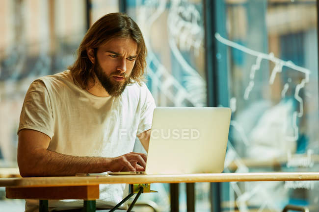 Joven hombre guapo barbudo sentado en la cafetería exterior y trabajando con el ordenador portátil en la mesa - foto de stock