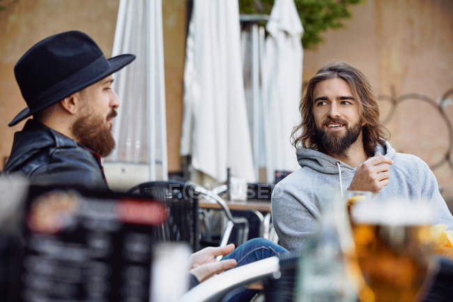 Красивый мужчина в черной шляпе сидит и наслаждается беседой с другом в серой толстовке на улице кафе — стоковое фото