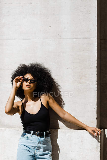 Чувственная этническая женщина в джинсах и майке, опирающаяся на стену и смотрящая на камеру на открытом воздухе — стоковое фото