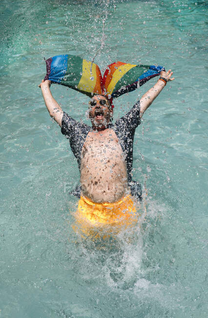 Fröhlicher Mann in Badebekleidung schreit und schwenkt LGBT-Flagge beim Plantschen im Schwimmbad des Resorts — Stockfoto