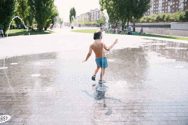 Back view of anonymous little boy in swimsuits standing near jet of water splashing out of fountain on street — Stock Photo