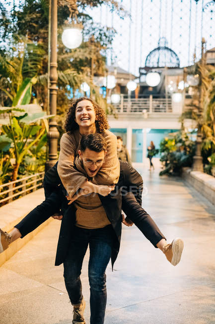 Aufgeregter junger Mann gibt Huckepack-Fahrt zu fröhlichen jungen Frau, während zu Fuß entlang des Weges bei romantischen Date auf der Straße der Stadt — Stockfoto