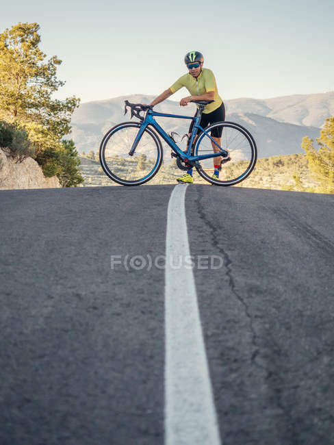 Здоровый человек отдыхает стоя с велосипедом на горной дороге в солнечный день — стоковое фото