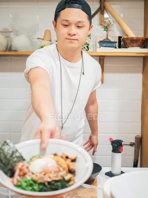 Jovem de pé na cozinha e tigela de mão com prato japonês cozido fresco — Fotografia de Stock