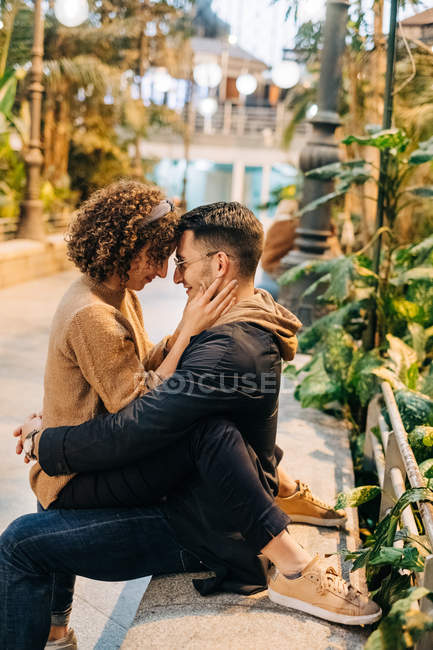 Vista lateral de los hombres y mujeres jóvenes abrazando y tocando la frente mientras están sentados en el banco durante una cita romántica en la calle de la ciudad - foto de stock