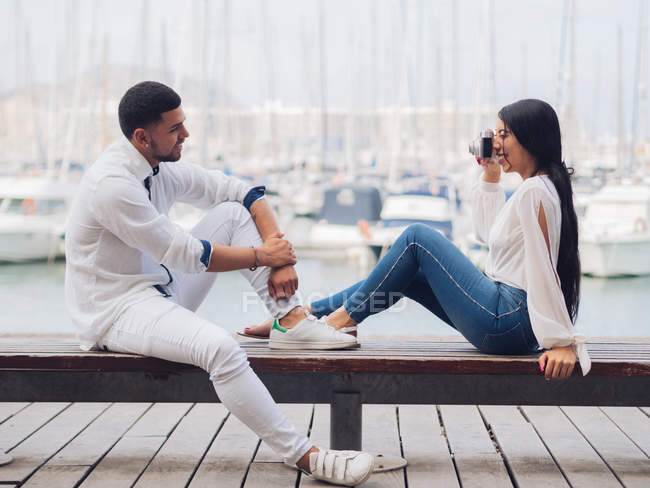 Side view of young couple sitting and having fun taking picture on wooden bench on sandy seaside — Stock Photo