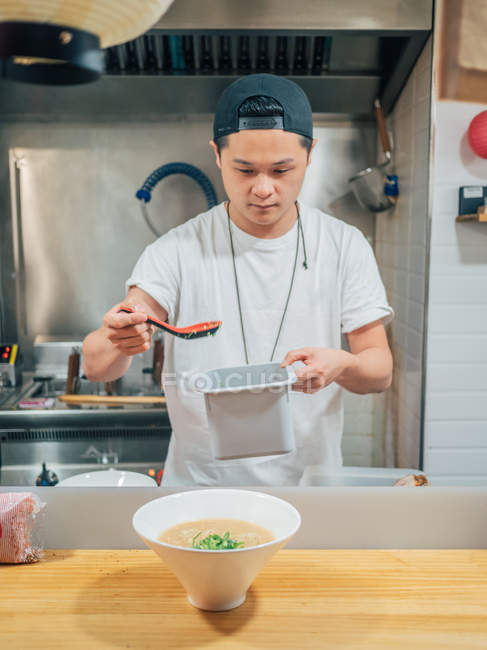 Asiatische Männchen Hinzufügen gehackter grüner Zwiebeln in Schüssel mit frisch gekochtem japanischem Gericht auf Holztisch — Stockfoto