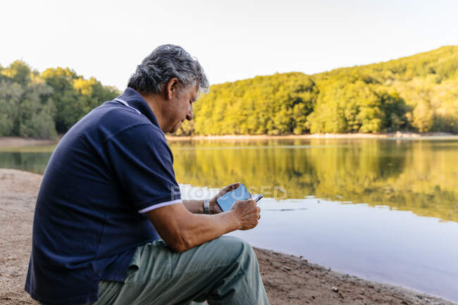 Вид сбоку пожилого человека, который пользуется мобильным телефоном, сидя на удивительном озерном горном ландшафте — стоковое фото