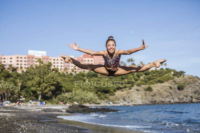 Schöne flexible Frau springt mit ausgestreckten Beinen und Armen und schaut in die Kamera am Strand — Stockfoto