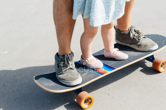 Безликий мужчина в кроссовках и маленькая девочка в платье, стоящие вместе на скейтборде — стоковое фото