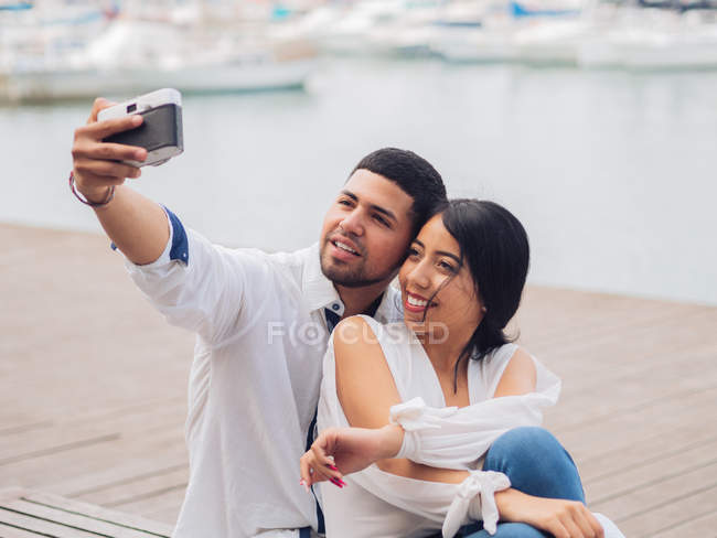 Young couple sitting and having fun taking a selfie picture on wooden bench on sandy seaside and kissing — Stock Photo