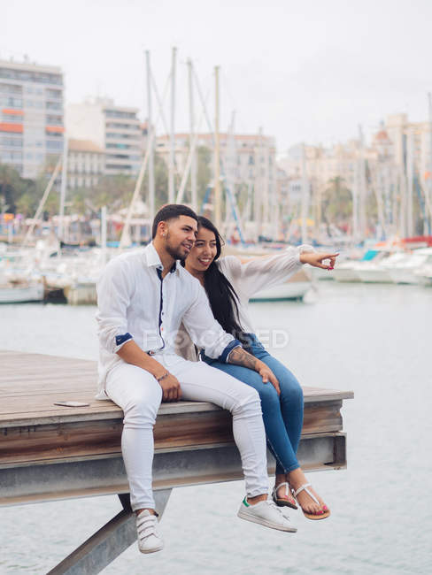 Young lovely couple sitting on dock with feet and embracing looking along — Stock Photo