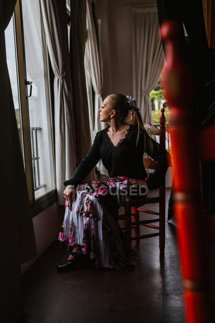 Привлекательная вдумчивая танцовщица в цветочной юбке для сидения фламенко и выглядывания в окно — стоковое фото