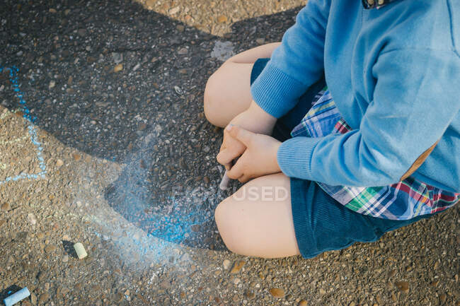 From above drawing with colorful chalks kid in bright blue clothes sitting on asphalt with crossed legs in sunny day — Stock Photo