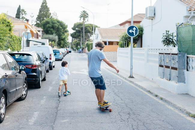 Back view of man on skateboard and boy on scooter riding down city street together — Stock Photo