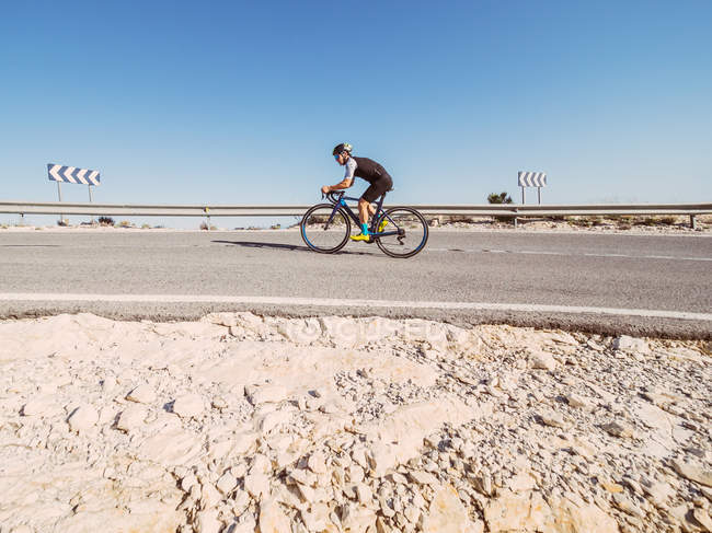 Homem saudável andar de bicicleta na estrada em dia ensolarado com paisagem estéril no fundo — Fotografia de Stock