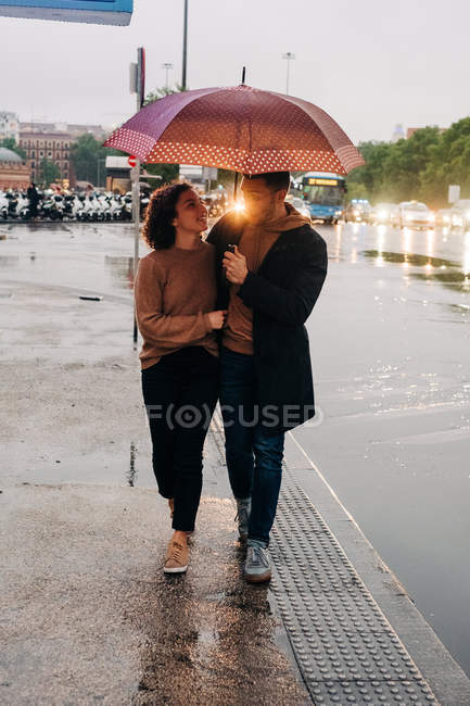 Fröhlicher junger Mann und Frau mit Regenschirm, die sich umarmen und einander anschauen, während sie an einem regnerischen Tag auf der nassen Stadtstraße stehen — Stockfoto
