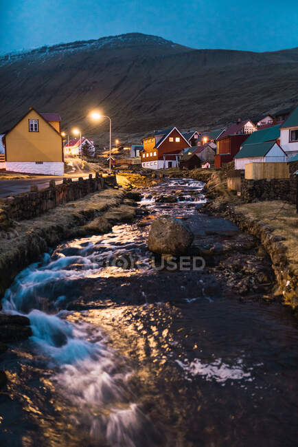 Pequeno riacho com água limpa perto de assentamento iluminado contra encosta à noite nas Ilhas Faroé — Fotografia de Stock
