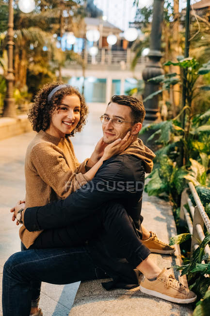 Vista lateral de hombre joven y mujer abrazando y mirando a la cámara mientras está sentado en el banco durante la cita romántica en la calle de la ciudad - foto de stock