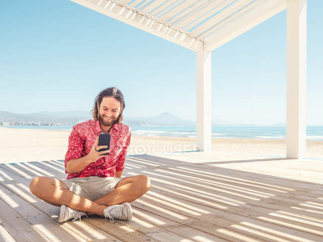 Homem barbudo navegando smartphone enquanto sentado no gazebo na praia de areia perto do mar no dia ensolarado — Fotografia de Stock
