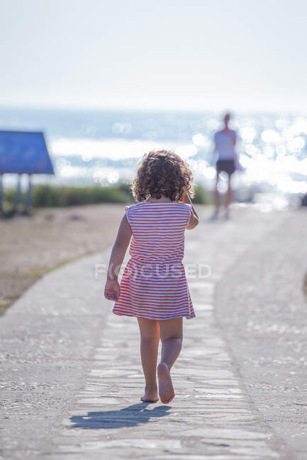 Вид на кудрявого ребенка в полосатом летнем платье, идущего вдоль дорожки на пляже на размытом фоне природы — стоковое фото