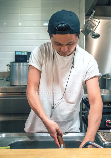 Hombre joven asiático en camiseta blanca y tapa negra cocinando plato de ramen japonés en la cocina - foto de stock