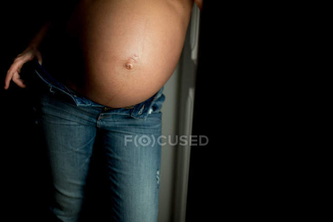 Брюхо анонимной беременной женщины в джинсах, стоящей у открытой двери дома — стоковое фото