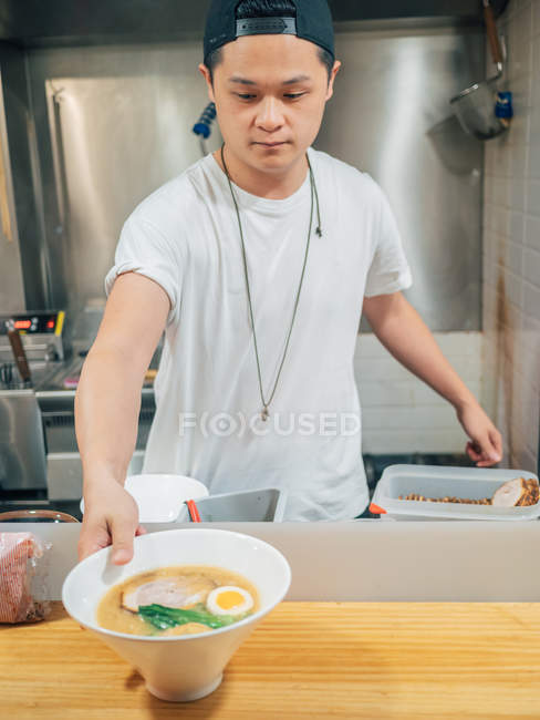 Asiatischer Mann legt Ei in Schüssel mit frisch gekochtem Ramen in Restaurantküche — Stockfoto