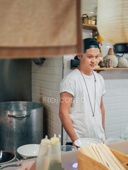 Jovem de pé com as mãos nos bolsos enquanto cozinha prato japonês ramen no restaurante — Fotografia de Stock