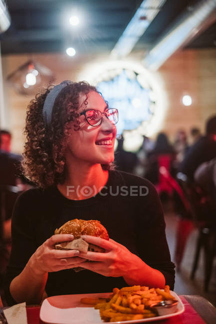 Mujer joven hambrienta comiendo sabrosa hamburguesa mientras está sentado en la mesa en la cafetería iluminada brillante - foto de stock