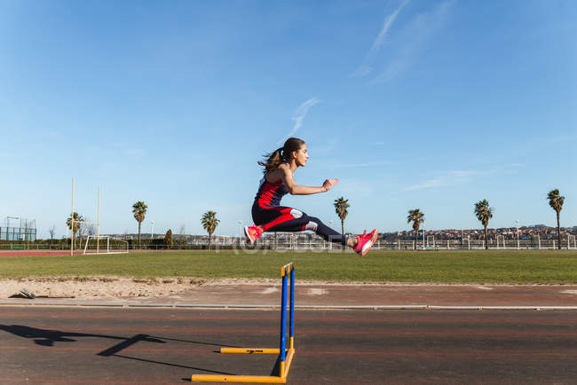 Сильна молода жінка в спортивному одязі стрибає через перешкоду на блакитному небі під час тренування на стадіоні — стокове фото
