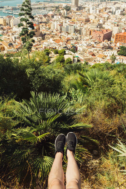 Von oben grüßen nackte Beine in schwarzen Turnschuhen hoch oben über üppigen Bäumen und dem Stadtbild in Alicante Spanien — Stockfoto