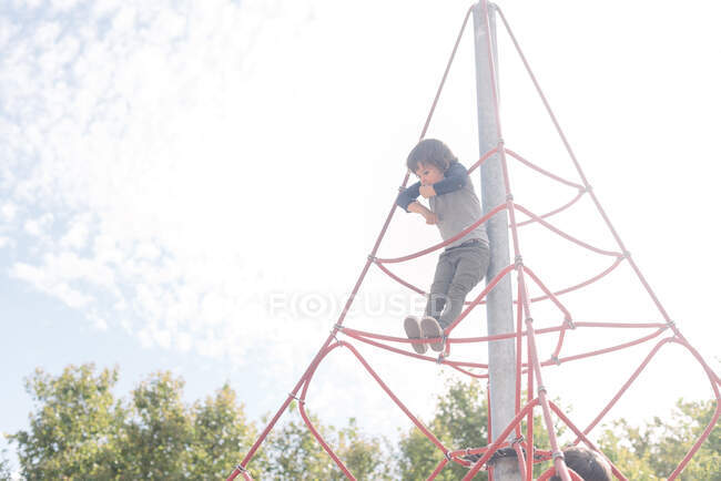 Pensive relaxed kid hanging on rope climbing net on playground in bright light — Stock Photo
