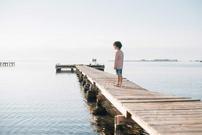 Вид сзади на маленького мальчика, стоящего на длинном деревянном пирсе, мечтательно смотрящего под солнечным светом — стоковое фото