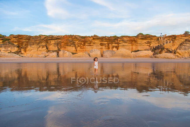 Кудрявый ребенок ходит по воде по песчаному пляжу на размытом природном фоне — стоковое фото