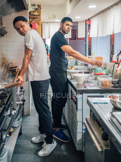 Vista lateral de los hombres jóvenes multirraciales cocinar plato japonés llamado ramen en el restaurante asiático y mirando en cámara - foto de stock