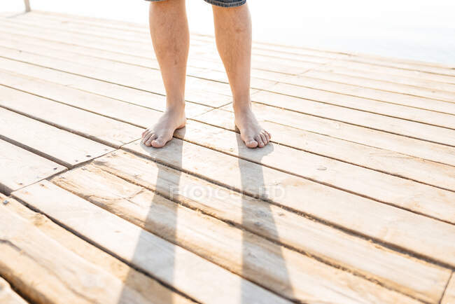 Vista de la cosecha de las piernas del hombre adulto de pie descalzo en el muelle de madera en el sol - foto de stock