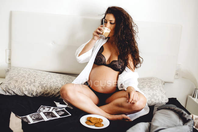 Pregnant woman in underwear sitting cross-legged with ultrasound scans while having snack in bedroom — Stock Photo
