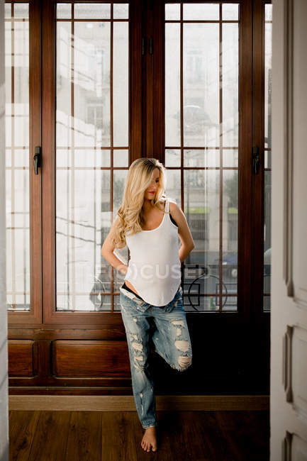 Задумчивая беременная женщина в повседневной одежде стоит у двери дома — стоковое фото
