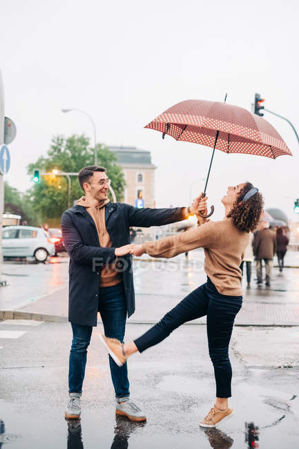 Весёлый молодой человек и женщина с зонтиком обнимаются и смотрят друг на друга, стоя на улице в дождливый день — стоковое фото