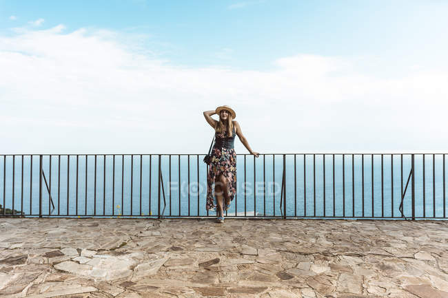 Frau in Sommerkleid und Strohhut steht am Zaun auf Steinterrasse mit Meereslandschaft im Hintergrund — Stockfoto