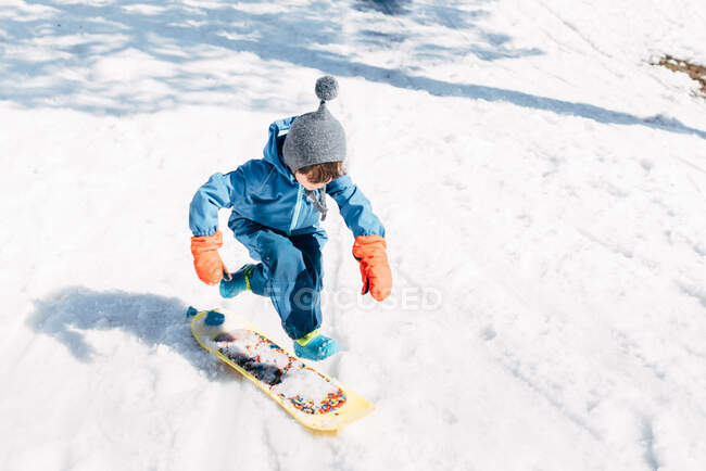 Garçon en vêtements d'hiver équitation petit conseil tout en glissant vers le bas montagne enneigée au soleil — Photo de stock