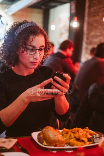 Mujer joven con el pelo rizado utilizando el teléfono inteligente para tomar una foto de hamburguesa y papas fritas mientras está sentado en la mesa de la cafetería - foto de stock