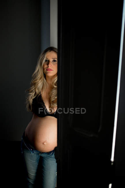 Attractive pregnant woman in bra looking at camera while standing near open door at home — Stock Photo