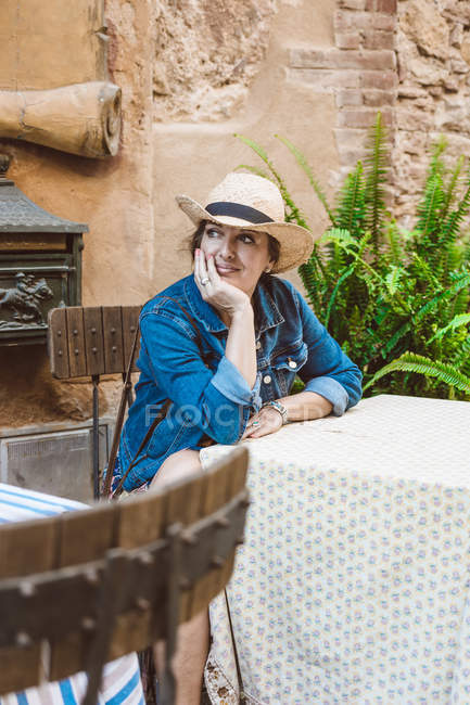 Женщина в соломенной шляпе и платье сидит на террасе ресторана в средневековом городе — стоковое фото
