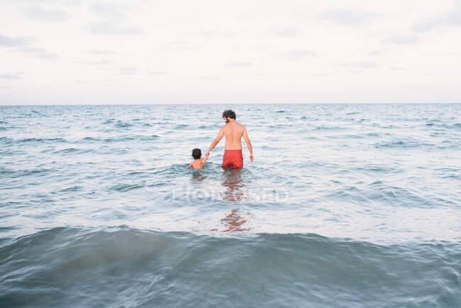 Visão traseira do homem com o menino de mãos dadas e andando na água nadando juntos — Fotografia de Stock