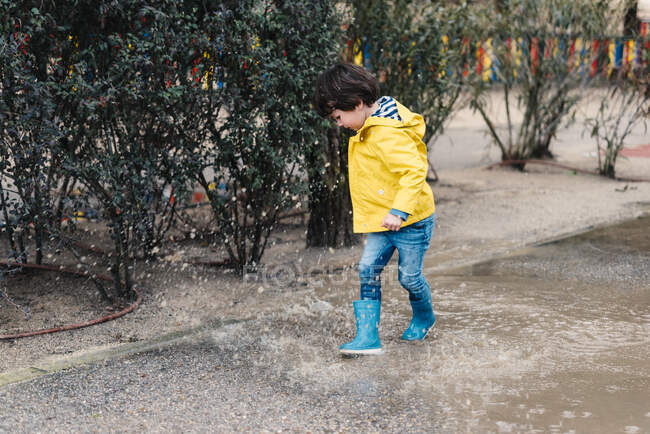 Excited boy in coat and gumboots having fun on street and jumping on puddle — Stock Photo