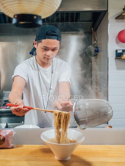 Молодой человек кладет горячую лапшу в миску палочками для еды во время приготовления японского блюда на кухне — стоковое фото
