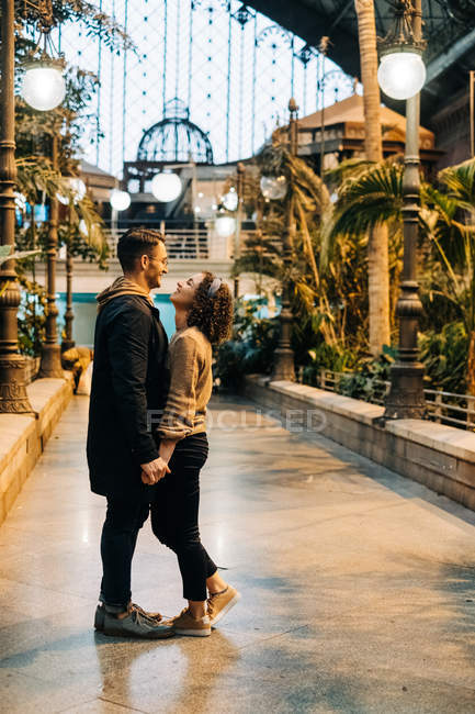 Cheerful young man and woman embracing and looking at each other while standing inside illuminated pavilion during date — Stock Photo