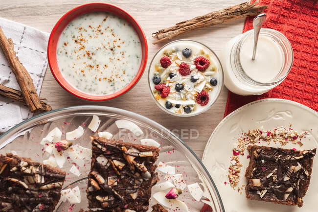 Von oben appetitlich duftender Joghurt mit Himbeer-Johannisbeerhafer im Glas und roten Schalen mit Zimtstangen und geschnittenem braunen Kuchen auf hölzernem Hintergrund — Stockfoto